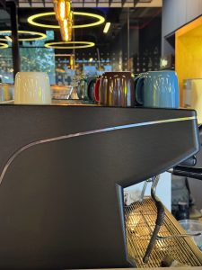 Close-up indoor shot of a professional espresso machine on a cafe counter, showing its dark metallic body and a grid drip tray, with colorful mugs lined up on top and warm ambient lighting overhead.   