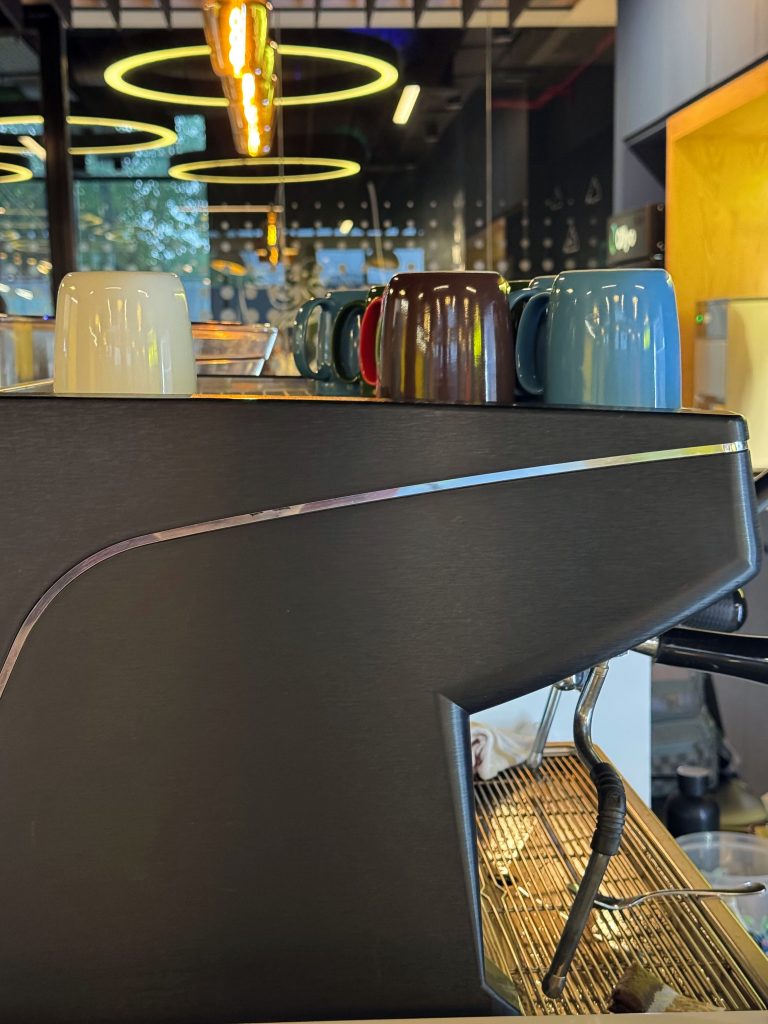 Close-up indoor shot of a professional espresso machine on a cafe counter, showing its dark metallic body and a grid drip tray, with colorful mugs lined up on top and warm ambient lighting overhead.