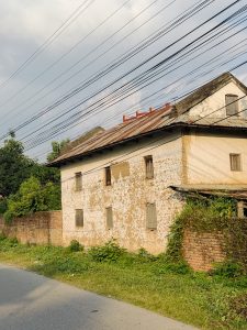 Weathered two-story building with peeling white paint, overgrown greenery, and power lines along a narrow road.
