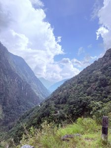 A scenic view looking down a lush green mountain valley flanked by steep, forested slopes under a sky filled with dramatic white clouds.