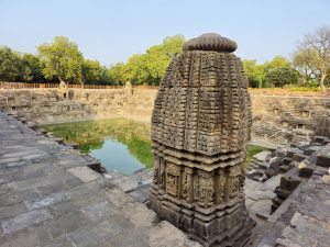 An intricately carved stone shrine stands beside the historic stepped tank at the Sun Temple. The green water, ancient steps, and surrounding trees create a beautiful composition. Location: Sun Temple, Modhera, Mehsana district, Gujarat.

