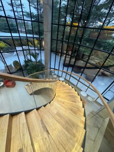 High-angle indoor shot looking down a graceful wooden spiral staircase with a clear glass railing and wooden handrail, leading past large multi-pane grid windows that reveal an outdoor courtyard with modern seating and lush green trees. 
