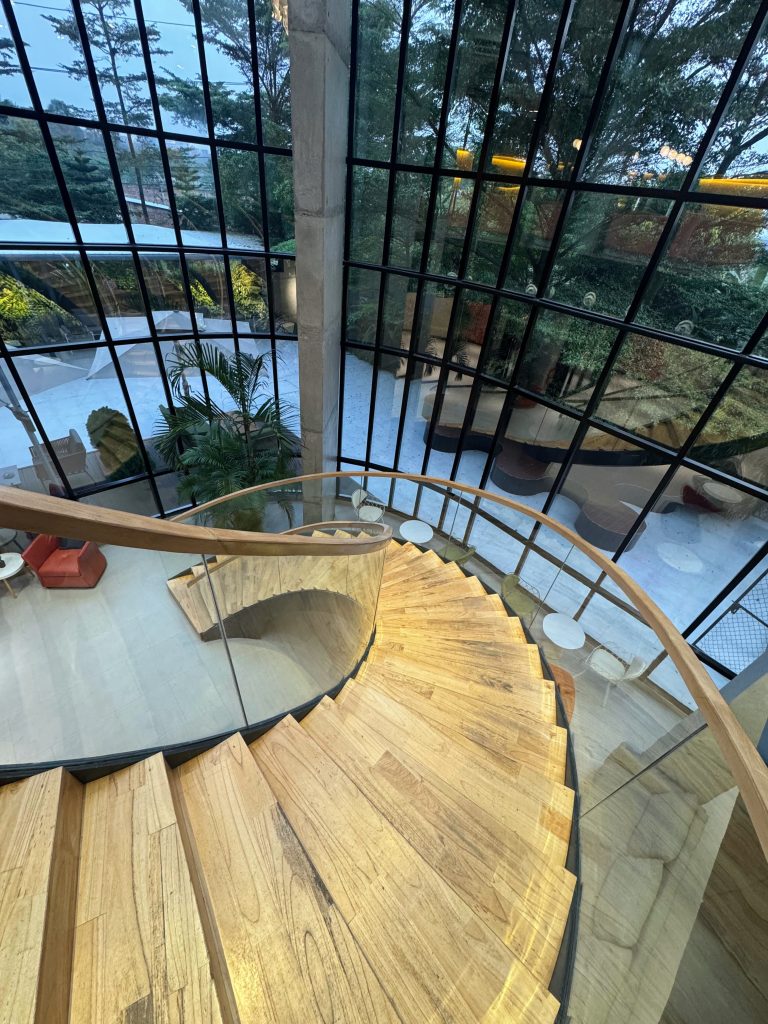 High-angle indoor shot looking down a graceful wooden spiral staircase with a clear glass railing and wooden handrail, leading past large multi-pane grid windows that reveal an outdoor courtyard with modern seating and lush green trees.