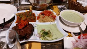 A platter of grilled meat, curry, seasoned rice, cucumber salad, and mint yogurt sauce served with a bowl of creamy soup and sliced onions.
