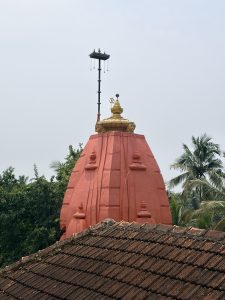 An eye level shot captures the upper portion of a traditional temple spire, painted in a bright reddish pink hue, rising above a weathered tiled rooftop in the foreground against a hazy sky.