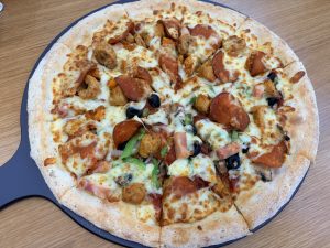 A large, round pizza with a golden crust, loaded with melted mozzarella cheese, pepperoni slices, chicken pieces, green bell peppers, black olives, and mushrooms, served on a black pizza peel.