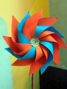Close-up shot of a colorful handmade paper pinwheel with alternating orange and blue curved blades mounted on a thin stick, set against a blurred warm-toned background.