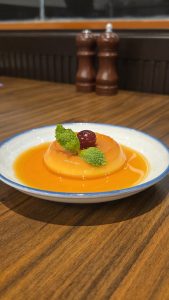 A classic caramel custard pudding served on a white plate with a pool of amber caramel sauce, garnished with a fresh cherry and mint leaves.