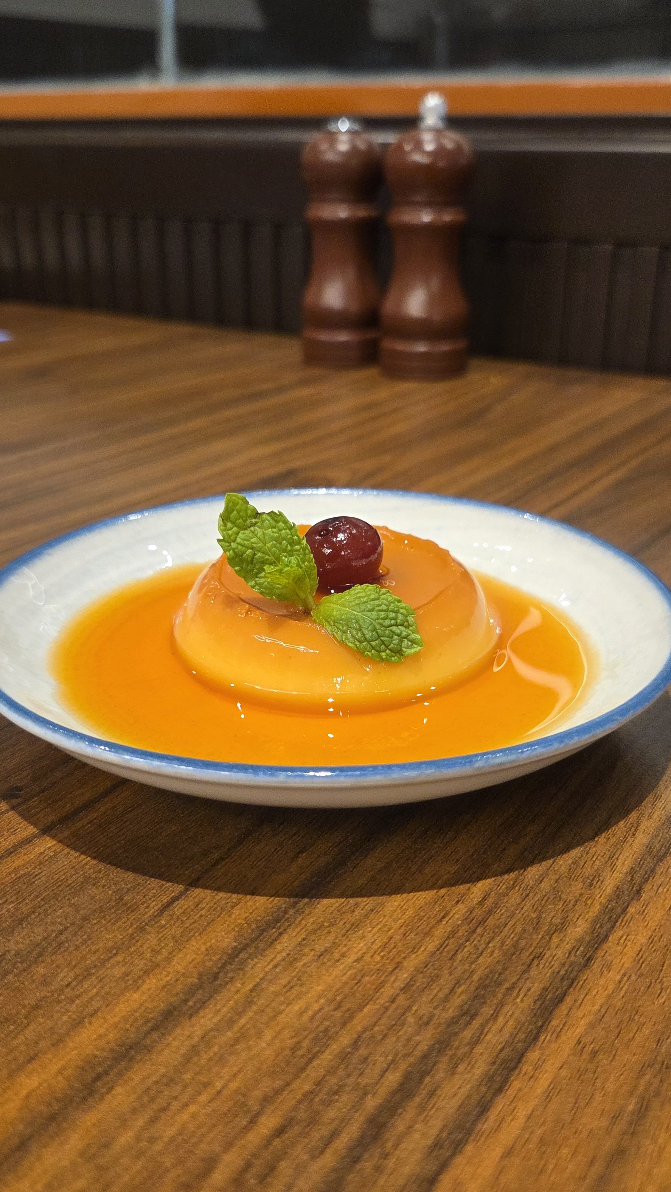 A classic caramel custard pudding served on a white plate with a pool of amber caramel sauce, garnished with a fresh cherry and mint leaves.