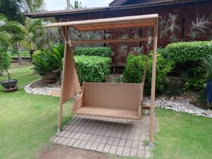 A brown woven wicker outdoor canopy swing bench placed on a stone tile pad on a green lawn surrounded by shrubs.