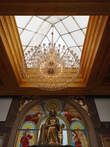 A grand crystal chandelier suspended beneath a glass ceiling and an ornate gold frame, positioned directly above a seated bronze statue flanked by vibrant stained-glass arch windows.