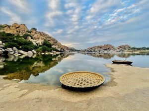 A traditional round coracle rests on a sandy granite riverbank beside calm water. Rocky hills, green vegetation, and scattered clouds reflect across the river, capturing the natural beauty of evening time and traditional river transport of the region. Location: Hampi, Karnataka.