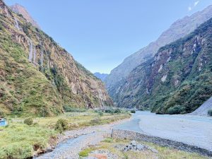 A wide view of a turquoise river flowing through a deep mountain valley with steep, green slopes and small waterfalls cascading down rock faces under a pale blue sky.