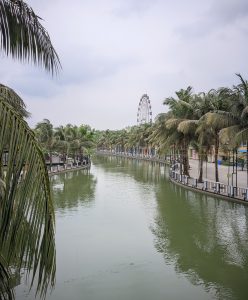 A calm canal winding through an amusement park, lined with palm trees and fenced walkways, with a Ferris wheel visible in the distance beneath a cloudy sky.
