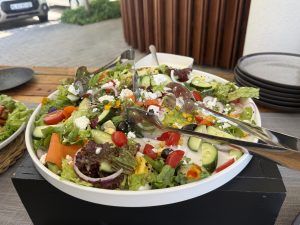 A vibrant salad in a white bowl contains mixed greens, cherry tomatoes, cucumbers, avocado slices, corn, red onions placed on a wooden outdoor table, suggesting a fresh and inviting meal.