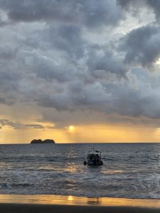 Sunset over the sea with a small boat, distant island, and golden light reflecting on the water.
