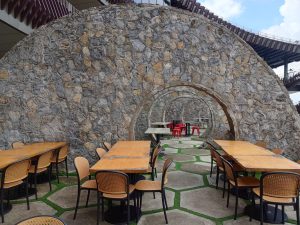 An outdoor dining area with wooden tables and woven chairs set around a series of concentric archways built from stone masonry.