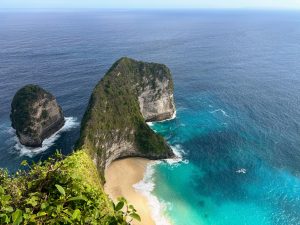 Nusa Penida beach with turquoise water, green cliffs, and a rocky islet offshore.