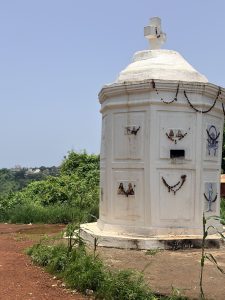 White roadside shrine with a stone cross, religious pictures, and decorative chains, surrounded by greenery with a hillside village in the background.