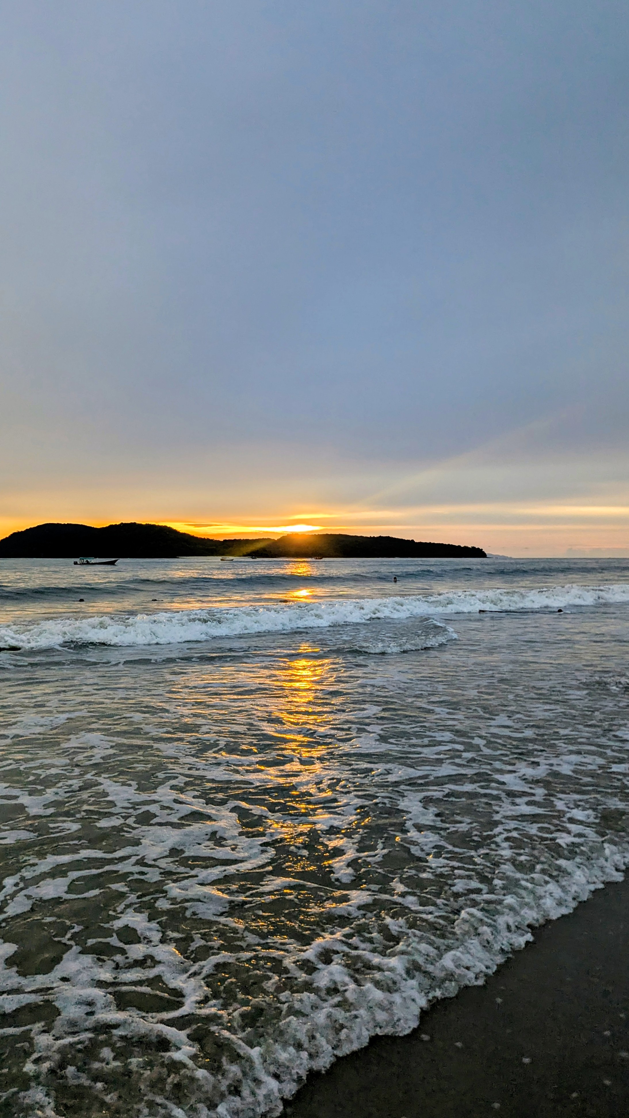 Sunset over the ocean with gentle waves reaching the shore and a silhouette of an island in the distance under a cloudy sky.