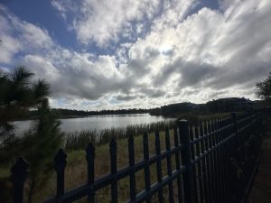 A serene outdoor scene featuring a calm lake bordered by tall grasses and a fence. The view includes a blue sky partially covered by fluffy white and grey clouds. There are hints of sunlight peeking through the clouds, creating a soft ambiance. In the background, there are some buildings that appear to be residential, set back from the water's edge