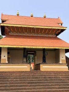 A traditional Kerala-style temple building with a two tiered bright orange tiled roof, wooden pillars, and large stone steps leading up to the entrance under a clear blue sky.