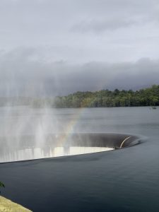 A large circular water spillway built into a misty reservoir under an overcast sky. Water rushes over the curved edge into the massive open sinkhole. A vibrant rainbow arcs through the rising mist directly over the spillway opening, while dense green trees and hills line the distant shore. Salaulim Dam located in South Goa, India.