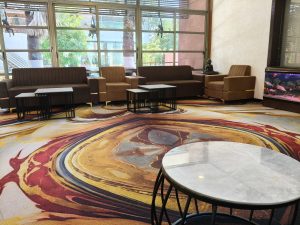 Comfortable lounge with brown sofas, an abstract colorful carpet, tables, a large window, and a lit aquarium.