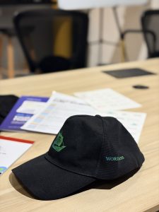 A black cap sits on a light wood conference table during the WP Future Conclave 2026. The front of the cap features a green embroidered logo of a stylized sailing ship containing a WordPress Logo on its sail, while the side is embroidered with teal text reading "WORDPRESS ".