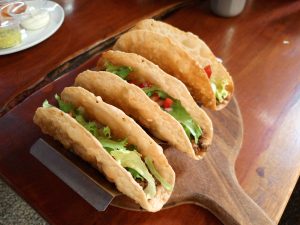 Three crispy hard-shell tacos filled with seasoned minced meat, shredded green lettuce, and diced tomatoes arranged on a wooden serving board.