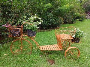 A copper-painted wire tricycle planter in a garden displaying potted blooming flowers across its front basket and rear passenger frame on green grass.
