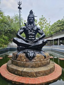 A magnificent black statue of Lord Shiva seated in meditation on a sculpted stone pedestal surrounded by a pond. The trident, lush greenery, and temple surroundings create a peaceful atmosphere at Ayamkudy, Kottayam, Kerala.
