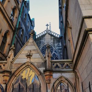 Looking up at a Gothic Revival stone church squeezed between buildings: pointed arches, carved pinnacles, a blue stained-glass rose window, and a cross on the roof, with black iron fence spikes in the foreground.