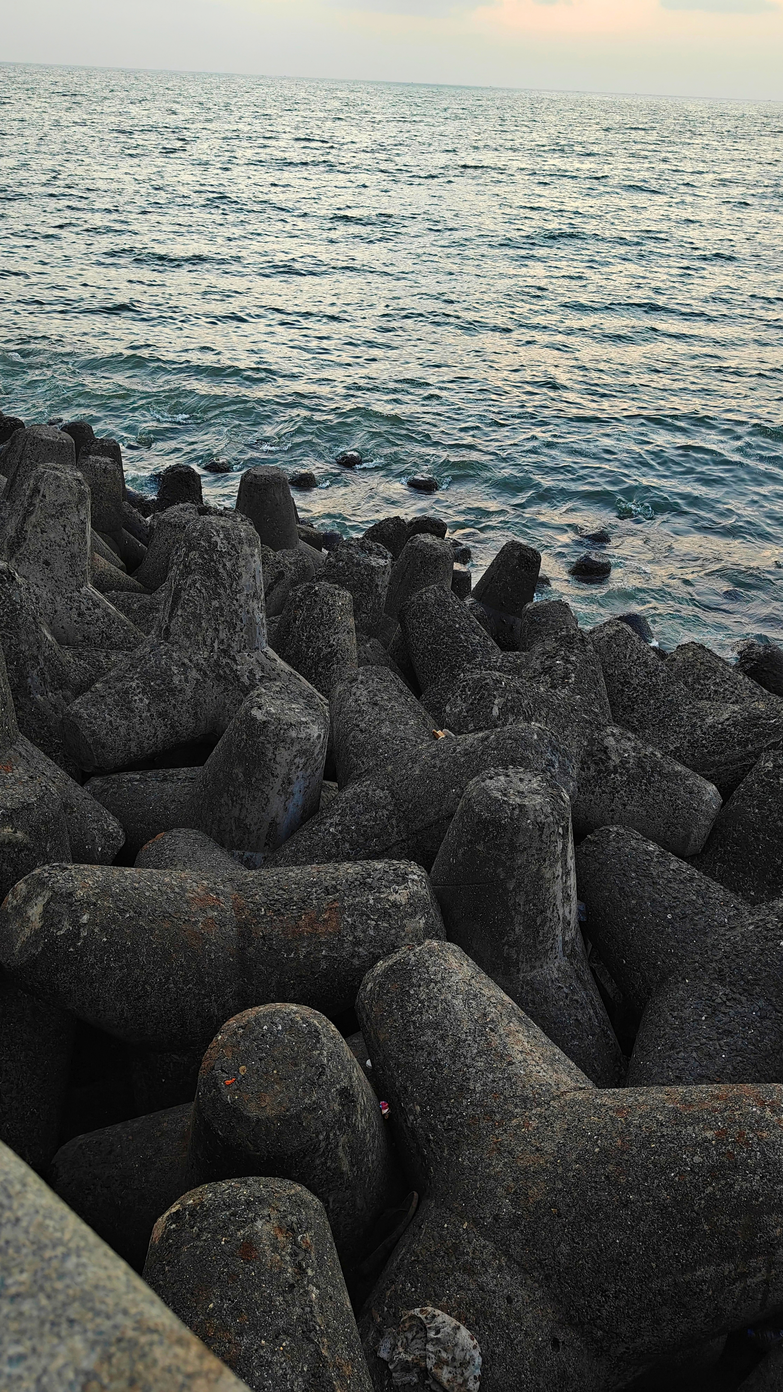 A scenic coastal marine drive Mumbai view featuring dark, rugged concrete wave breakers along the shoreline, with gentle waves rippling across the blue-green sea beneath a pale, hazy sky.