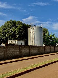 A large silver water tank stands behind a concrete wall with trees and banana plants around it under a blue sky.