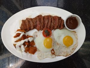 A white plate containing sliced grilled beef steak, two sunny-side-up fried eggs, roasted potato cubes drizzled with mayonnaise, a grilled tomato slice, and a cup of steak sauce.