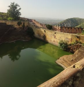 Historic fort walls surrounding a green water reservoir, with the city of Jaipur and hills visible in the distance.
