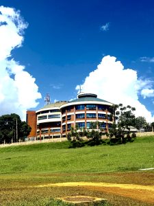 A round multi-storey brick building on a grassy hill under a bright blue sky with white clouds.
