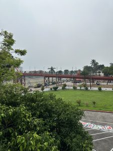 A green lawn with shrubs in the foreground, a red pedestrian bridge, construction vehicles, and palm trees under a cloudy sky in the background. 
