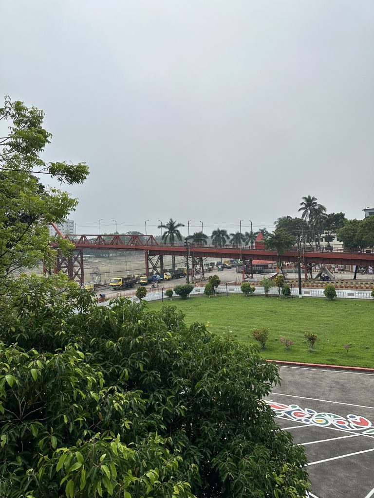 A green lawn with shrubs in the foreground, a red pedestrian bridge, construction vehicles, and palm trees under a cloudy sky in the background.