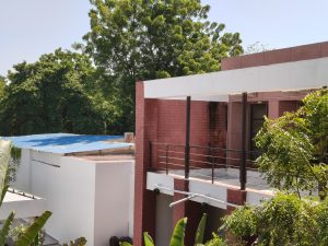Modern red-and-white building with an outdoor balcony, metal railings, and surrounding green trees under a clear sky.