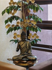 Bronze Buddha statue seated among leaves and golden flowers, with cushions on either side.
