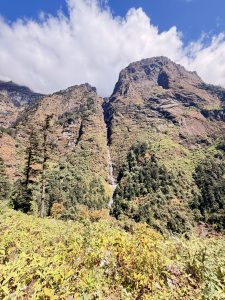 A dramatic, narrow waterfall cascading down a towering, sheer rock face nestled between steep, forested mountain ridges under a partly cloudy blue sky.