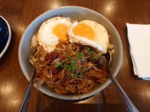 A Filipino cuisine rice bowl featuring shredded savory chicken adobo topped with chopped scallions and two sunny-side-up fried eggs.