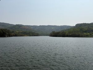 A calm lake surrounded by lush green hills and dense forests under a clear, hazy sky.