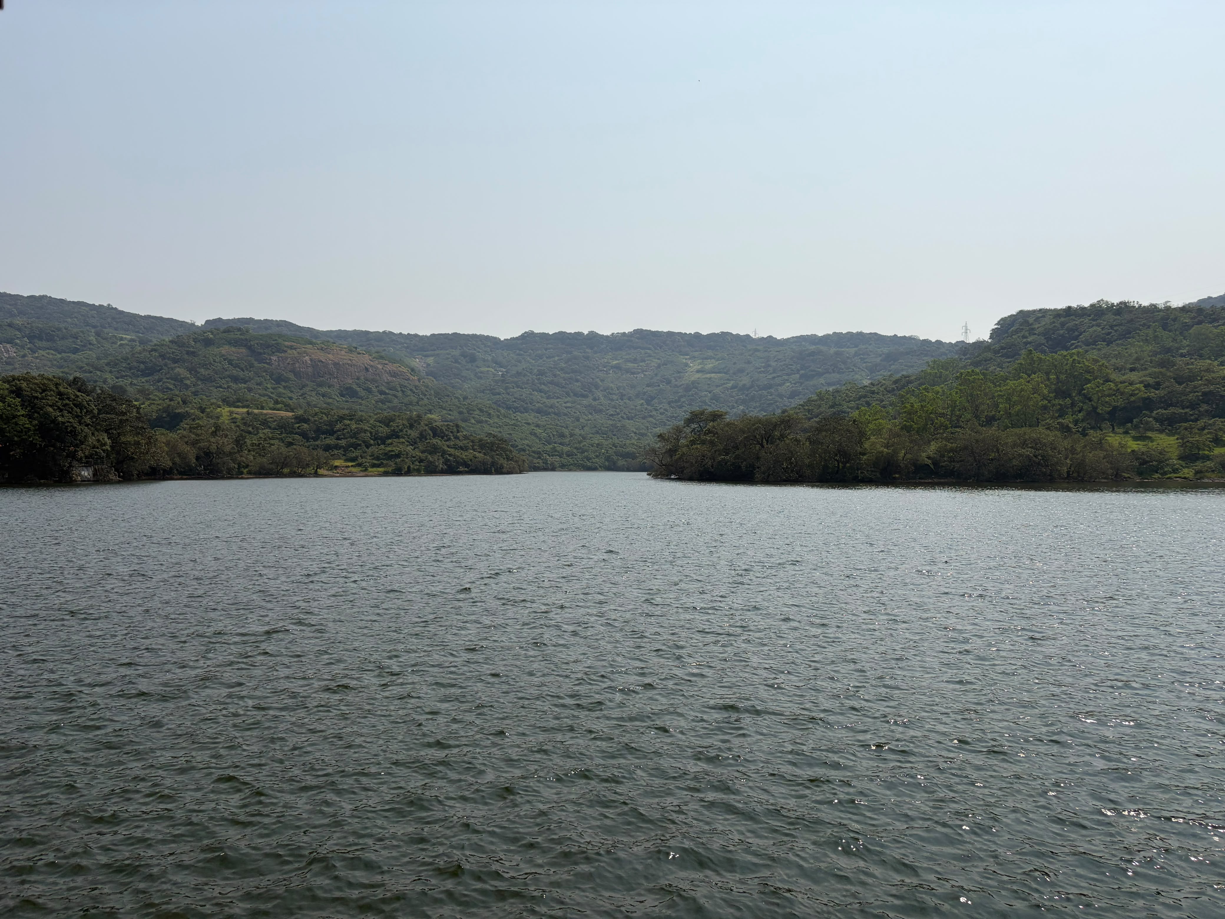 A calm lake surrounded by lush green hills and dense forests under a clear, hazy sky.