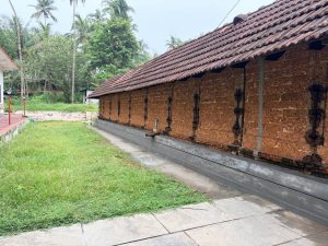 A long, traditional Kerala-style temple building features a weathered, sloped roof covered in reddish-brown clay tiles. The exterior wall is constructed from exposed, rough-textured red laterite stone blocks, punctuated by narrow vertical wooden windows with black iron grills spaced evenly along the side.