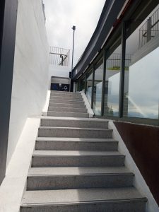 A flight of outdoor stone stairs leading up to a rooftop flanked by a white plastered wall on one side and a modern glass window on the other.