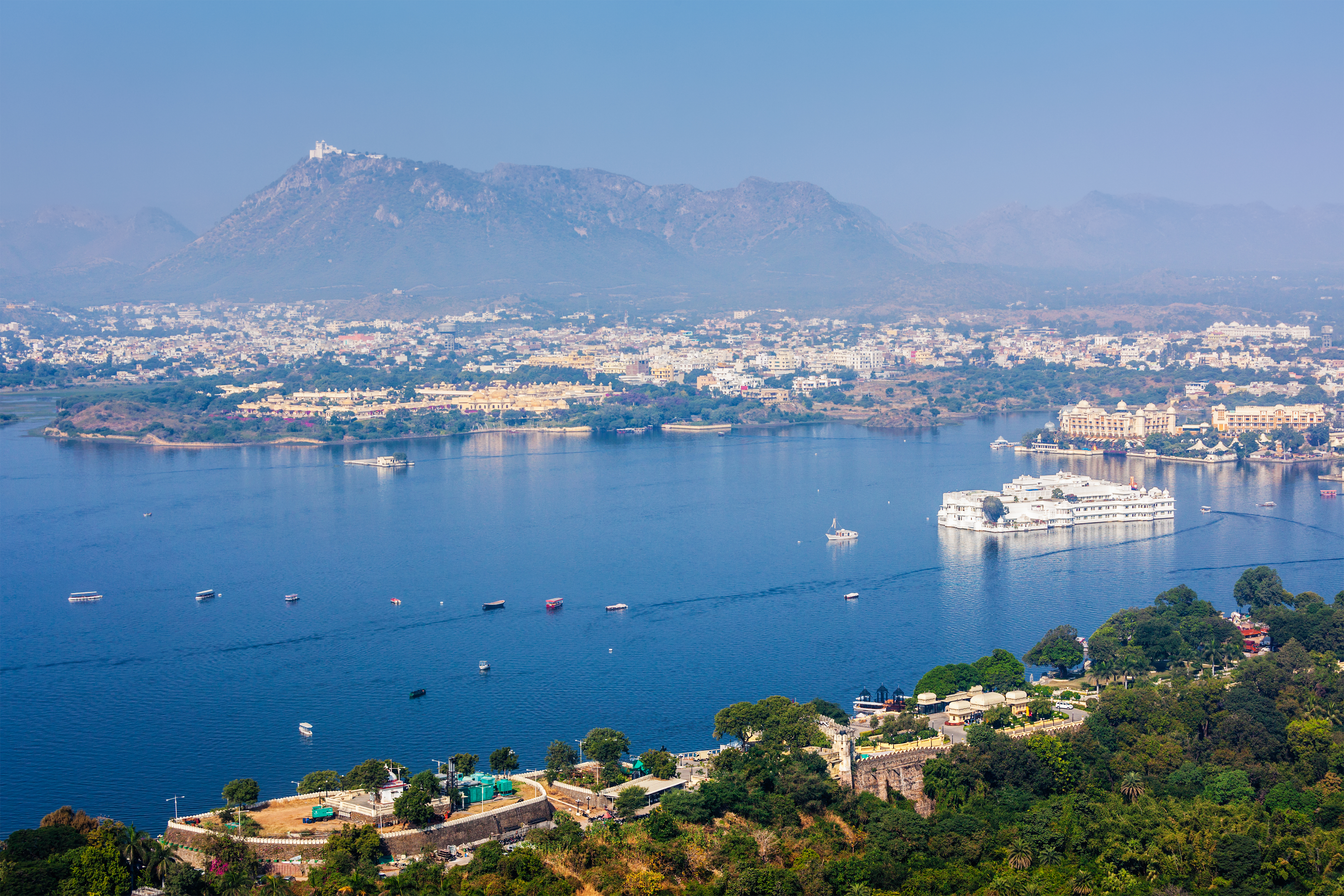 Lake Pichola Aerial View: A panoramic view of Lake Pichola in Udaipur, surrounded by the city and Aravalli hills. Boats dot the blue water, while the white Lake Palace and historic buildings appear along the shoreline.