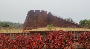 
A large stone observation tower and stairway at Bekal Fort in Kerala, surrounded by fort walls, green lawns, and red-leafed shrubs under a cloudy sky.